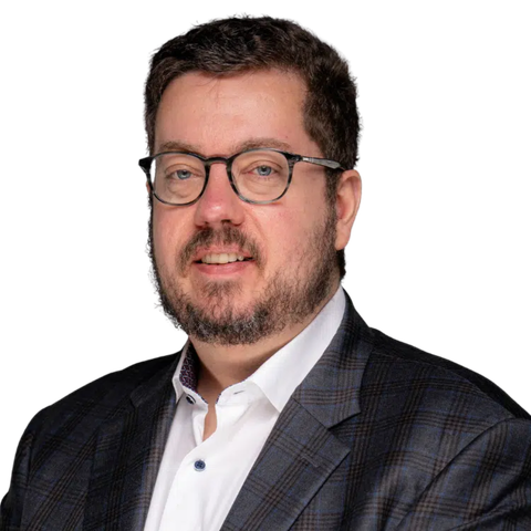 A man with short dark hair, glasses, and a beard, wearing a white shirt and a dark plaid blazer, smiling at the camera against a plain light background. EFESO Management Consultants